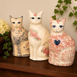 May include: Three ceramic cat figurines with floral designs. The cats are in cream, beige, and pink, with blue and pink floral patterns. One cat has a blue heart on its chest. The figurines are sitting on a dark wooden surface.