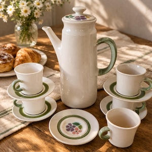May include: A white ceramic coffee pot with a lid featuring a floral motif, accompanied by four matching cups and saucers. Each piece has a green painted handle and a green circular band on the saucer, with small purple and pink flower accents.
