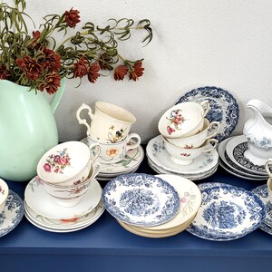 May include: A collection of vintage teacups, saucers, and plates with floral and blue patterns. A light green pitcher holds a bouquet of red flowers. The tableware is arranged on a dark blue surface, creating a classic tea party aesthetic.