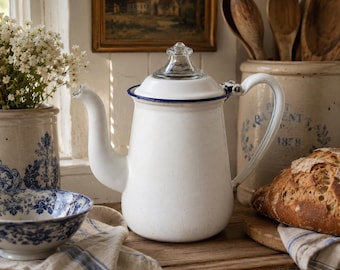 Puede incluir: Una cafetera de esmalte blanco con borde azul y tapa de cristal transparente. La cafetera presenta un pico curvo y un asa resistente. Es un recipiente de cocina clásico diseñado para preparar o servir café, con un acabado liso.