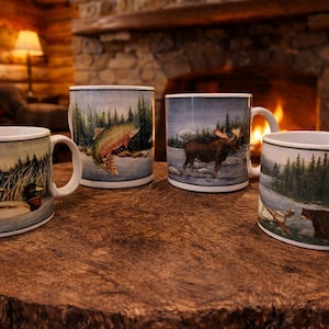May include: Four ceramic mugs with nature scenes, including a fish, moose, bear, and ducks. The mugs are white with a black rim and handle, set on a rustic wooden surface. A fireplace and lamp are in the background.
