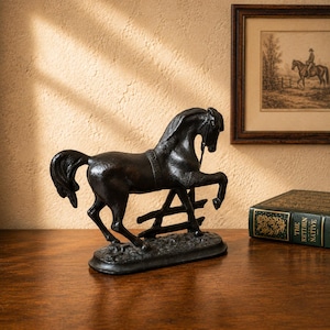 May include: A black cast iron horse figurine standing on an oval base. The horse is depicted in a dynamic pose with one front leg raised, positioned next to a small fence structure. The surface of the sculpture has a textured, matte finish.