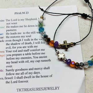 May include: Two black string bracelets with colorful beads and silver and gold cross charms. The bracelets are displayed on a white card with the text "PSALM 23" and a verse from the Bible.