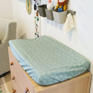 May include: A pale green changing mat with a white leaf design sits on a light wood chest of drawers. A stuffed lion toy, a bottle of lotion, and storage bins are mounted on the wall above the chest. A height chart hangs on the wall.