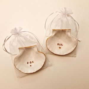 以下が含まれることがあります： 金色の縁取りが付いた、白い貝殻型の陶器製の小物入れが2つ。それぞれ透明な白い巾着袋に入っています。「ZOE」と星、「BEL」とハートの文字がそれぞれ書かれています。