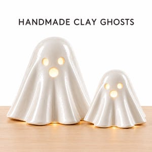 Pode incluir: Duas figuras de fantasmas em argila branca brilhante com olhos e bocas recortados, iluminadas por dentro por uma luz quente. O texto HANDMADE CLAY GHOSTS está impresso acima. Os fantasmas têm uma aparência lisa e drapeada.