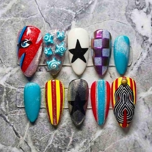 May include: A set of ten almond-shaped press-on nails featuring diverse designs including a red lightning bolt, blue swirls, black stars, purple checkered patterns, solid blue, yellow and red stripes, glitter, and black and white geometric lines.