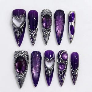 May include: A set of ten stiletto-shaped press-on nails in deep purple with metallic silver accents. Designs include raised silver heart motifs, large purple faceted gems, intricate silver filigree, textured leaf patterns, and glossy purple finishes.