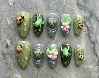 May include: A set of ten almond-shaped artificial nails with a nature theme. Each nail features a different design, including ladybugs, frogs, and lily pads. The nails are painted in shades of green, gray, and pink, with water droplet details.