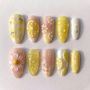 May include: A set of ten almond-shaped press-on nails featuring yellow and white designs. Patterns include yellow gingham checks, 3D lemon slices, raised white daisy flowers, clear water droplets, and small metallic studs on soft pink and yellow bases.