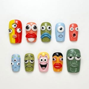May include: A set of ten press-on nails featuring various colorful, three-dimensional cartoon faces with raised eyes, mouths, and distinct expressions. Each nail has a unique design, including shades of yellow, red, green, blue, and pink.