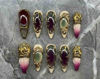 May include: A set of ten press-on nails featuring almond and stiletto shapes. The designs include deep purple and pale green centers encased in ornate gold filigree frames, alongside cream-colored tips with gradient magenta and gold swirl accents.