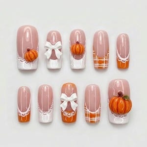 Peut inclure: Un ensemble de dix faux ongles avec une base nude et des accents orange et blancs. Les motifs incluent des citrouilles orange, des nœuds blancs, des bordures en dentelle blanche et des carreaux orange et blancs sur une forme allongée.