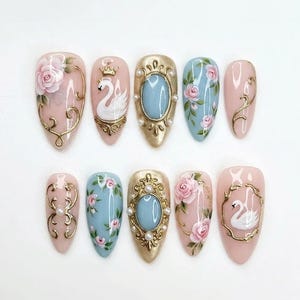 May include: A set of ten almond-shaped press-on nails featuring a variety of designs including hand-painted pink roses, white swans with gold crowns, light blue backgrounds, gold filigree accents, and small white pearls set in gold frames.