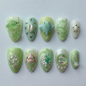 May include: A set of ten press-on nails in shades of light green and white, featuring 3D ocean-themed embellishments including a shark, stingray, turtle, seahorse, coral, seashells, pearls, bubbles, and the text SEA YOU on one nail.