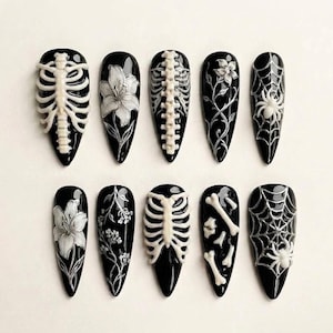 May include: A set of ten almond-shaped press-on nails with a glossy black base. The designs feature white raised 3D details including human rib cages, spines, lilies, intricate floral vines, spider webs with spiders, and scattered bone fragments.