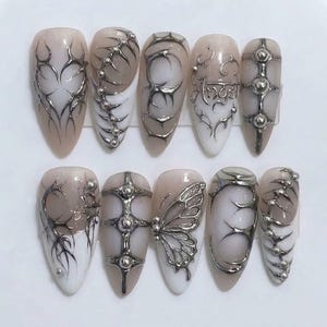May include: A set of ten almond-shaped press-on nails featuring a nude and white gradient base. The designs include silver metallic 3D accents, such as intricate tribal-style lines, raised spherical studs, a butterfly motif, and the word Angel.