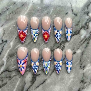 May include: A set of ten almond-shaped false nails. Each nail features a nude base with a colorful floral design in red, blue, yellow, and white. The designs are painted on the tips of the nails.