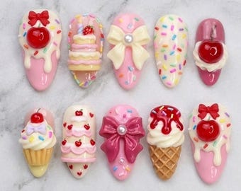 May include: A set of ten press-on nails featuring 3D decorations including miniature cakes, cupcakes, ice cream cones, cherries, bows with pearls, and colorful sprinkles. The designs are rendered in shades of pink, cream, red, and brown.