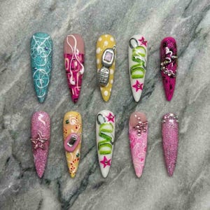 May include: A collection of ten long, pointed acrylic nails with various designs. The nails feature glitter, abstract art, and 3D embellishments, including a cell phone charm and the word "DVD". Colors include pink, blue, yellow, and white.