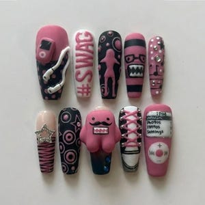 May include: A set of ten press-on nails featuring various designs in pink, black, and white. Patterns include a music player with headphones, the text #SWAG, a silhouette, a monster face, musical notes, a star, circles, and a sneaker lace design.