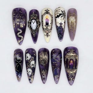 May include: A set of ten almond-shaped press-on nails featuring a Halloween theme with deep purple and cream backgrounds. Designs include a haunted house, a ghost in a frame, a lit candle, spiderwebs, skulls, a black cat, and a lantern.