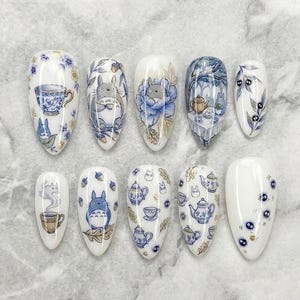 以下が含まれることがあります： 白い背景に青と金のティーセット、花柄、キャラクターのイラストが描かれた、アーモンド型のプレスオンネイル10本セット。デザインにはティーポット、ティーカップ、どんぐり、小さなススワタリが含まれています。