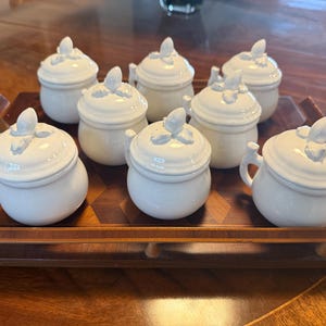 May include: Eight white ceramic lidded jars with handles, presented on a wooden tray. Each jar features a rounded shape and a floral motif on the lid. The tray has a dark wood finish and is rectangular.