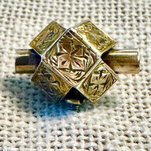 May include: A gold-toned metal charm with a geometric design. The charm is made up of multiple squares with intricate floral patterns. The charm has a cylindrical shape on either end.