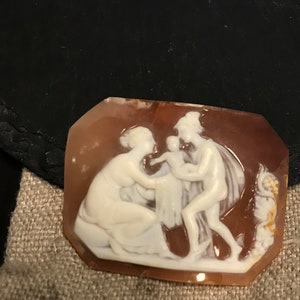 May include: A cameo carved from shell with a detailed depiction of three figures. The figures are carved in white against a brown background. The cameo is a rectangular shape with a beveled edge.