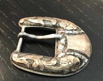 May include: A silver belt buckle with an ornate floral design. The buckle has a gold accent on the top and bottom.