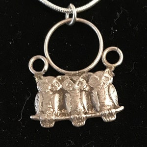 May include: A silver necklace with a pendant featuring four owls perched on a branch. The pendant has a circular hoop at the top.