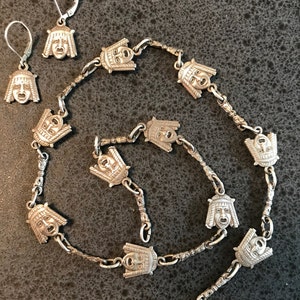 Könnte beinhalten: Eine silberne Halskette und Ohrringe mit einem sich wiederholenden Muster aus stilisierten Gesichtern. Die Halskette besteht aus einer Kette mit einer Reihe von silbernen Anhängern, die jeweils ein Gesicht mit einem dramatischen Ausdruck zeigen. Die Ohrringe sind einfache silberne Creolen mit einem Anhänger eines stilisierten Gesichts.