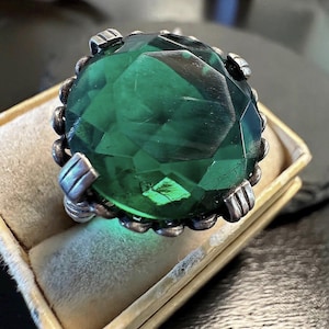 May include: A large, faceted emerald-green gemstone ring with a silver-toned setting. The ring is displayed in a beige velvet-lined box. The gemstone has a brilliant cut, reflecting light. The setting features decorative details.