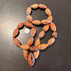 May include: A collection of intricately carved wooden beads, arranged in a figure-eight shape. The beads are a warm brown color and feature detailed carvings of human figures and other designs. A U.S. quarter coin is included for scale.