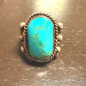 May include: A silver ring featuring a large, oval turquoise stone. The stone is a vibrant turquoise color with brown veining. The ring has a twisted rope design around the stone and small silver beads on the sides.