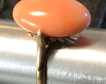 May include: A gold ring featuring a large, oval, peach-colored gemstone. The gemstone is set on a delicate, ornate gold band. The ring is displayed on a silver metal surface, showcasing its design and color.