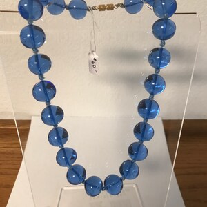 May include: A blue glass bead necklace with a gold clasp. The necklace is displayed on a clear stand. The tag on the necklace reads "4.00".