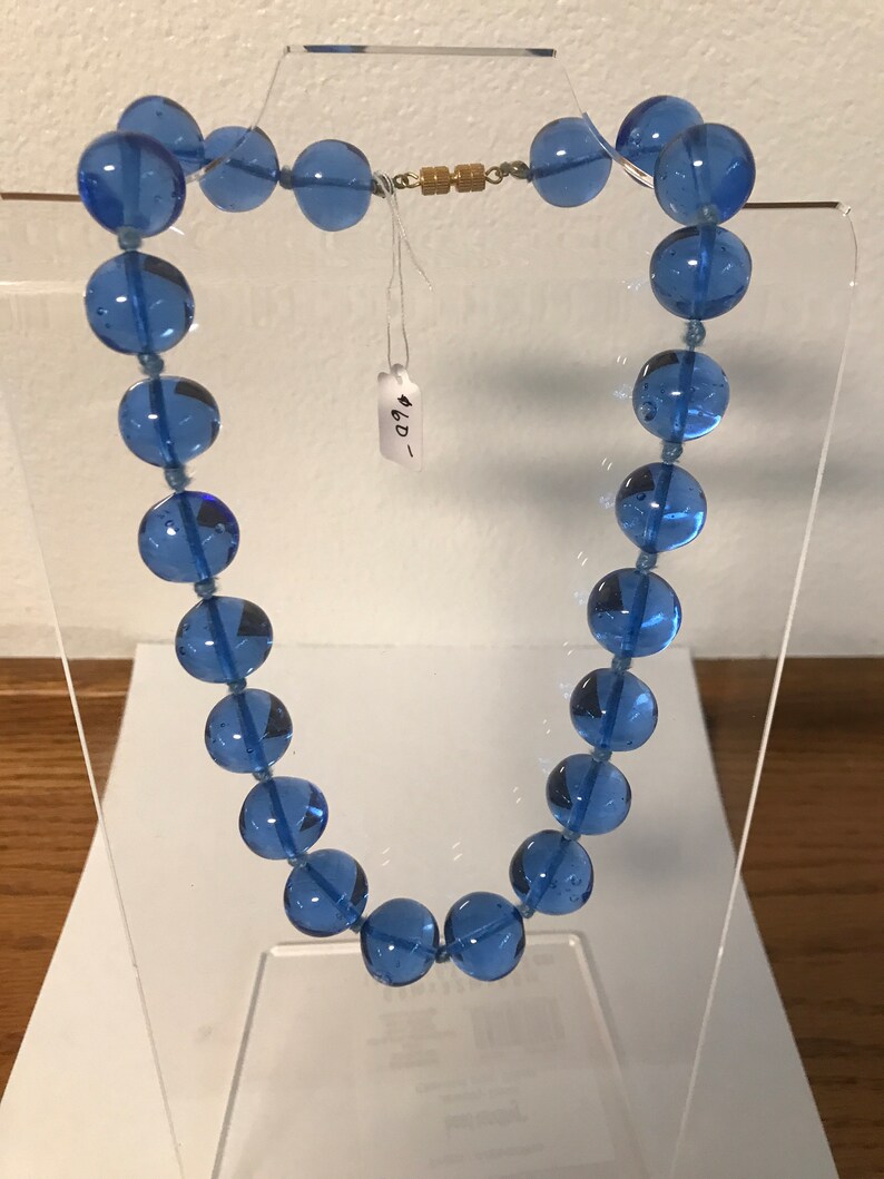 May include: A blue glass bead necklace with a gold clasp. The necklace is displayed on a clear stand. The tag on the necklace reads "4.00".
