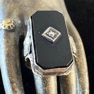 May include: A rectangular black onyx ring with a small diamond-shaped accent. The ring has a silver-colored band with floral and leaf detailing on the sides. The onyx is set in a silver bezel.