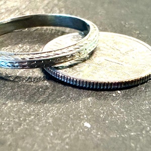 May include: A silver wedding band with a detailed engraved design, resting on a silver coin.