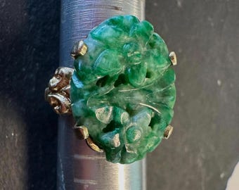 May include: A gold ring featuring a carved, oval-shaped jade stone. The jade is a vibrant green with intricate floral designs. The ring's band and setting are gold-colored, with small gold accents securing the jade.