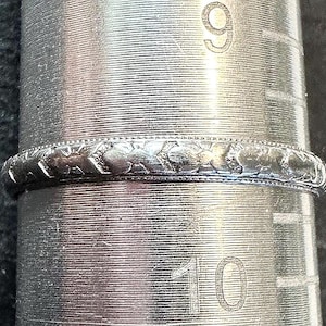 May include: A silver-toned metal ring with a repeating engraved pattern along the band, positioned on a metal ring mandrel that displays the number 9 and the number 10, indicating a size between 9 and 10 on the standard US ring sizing scale.
