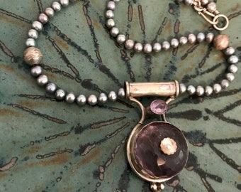 May include: A silver necklace with a large, round, faceted amethyst pendant. The pendant is surrounded by smaller amethyst stones and a silver flower design. The necklace is made of gray pearls.