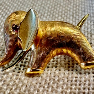 May include: A gold-toned elephant brooch with a red rhinestone eye. The elephant has a textured surface and a raised trunk.