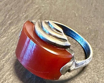 May include: A vintage ring featuring a rectangular, deep red stone set in a silver-toned metal band. The ring has a unique design with a stylized floral or leaf motif on the top and sides. The band is slightly tarnished, adding to its antique charm.