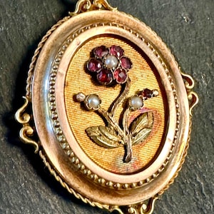 May include: A gold locket pendant featuring an intricate floral design. The flower is crafted from red and white gemstones and gold leaves. The locket is encircled by an ornate gold frame.