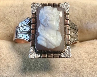 May include: A silver ring with a cameo set in a rectangular frame. The cameo features a white profile of a woman with a detailed hairstyle.