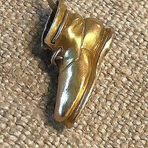 May include: A gold-colored pendant in the shape of a shoe, with a loop at the top for a chain. The shoe has a shiny, reflective surface and is set against a textured, neutral-toned background.