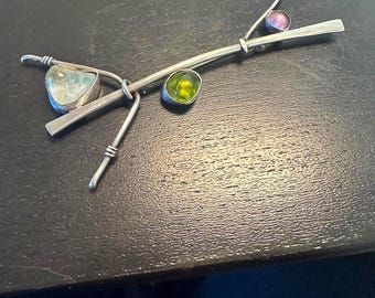 May include: A silver brooch with three gemstones: a light blue, a green, and a purple. The brooch is made of twisted wire and has a modern design.